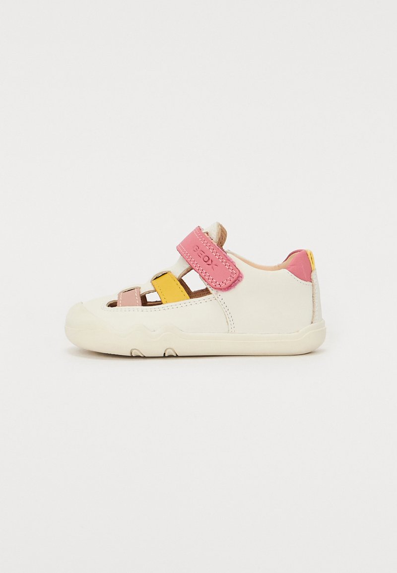 Sapatilha estilo ténis branca com acabamentos em rosa, amarelo e bege. Apresenta uma fita de velcro, laterais abertas e uma sola de borracha texturizada.