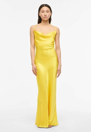 Kvinne stående iført en ermeløs, knallgul satengkjole med tynne stropper mot en ensfarget hvit bakgrunn.