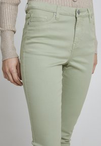 Jean taille haute vert clair avec une texture lisse, dotés de poches avant et de quincaillerie en ton argent pour la fermeture et les détails.