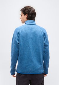 Νεαρός άνδρας με κοντά καστανά μαλλιά φοράει μπλε φλις μπουφάν με ψηλό γιακά, απεικονιζόμενος από πίσω σε απλό λευκό φόντο.