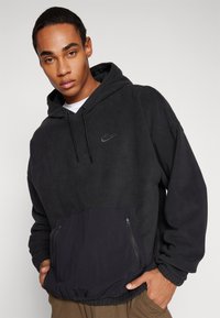 Črna flis bunda s kengurujskim žepom, kapuco s vrvico in logotipom na prsih. Ima kontrastni temni material žepa in elastične manšete.