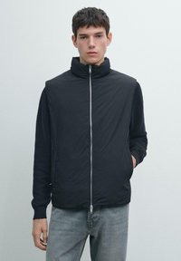 Czarna pikowana kamizelka z wysokim kołnierzem, zamkiem błyskawicznym z przodu i dzianinowymi rękawami. Nosi się ją na szare dżinsowe spodnie. Minimalistyczny design, gładka tekstura.
