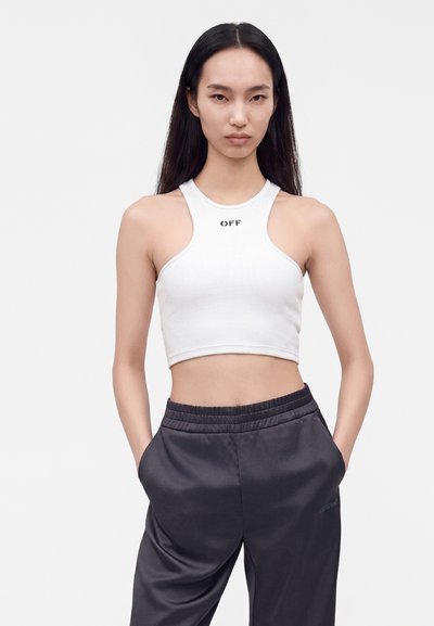Femme aux longs cheveux noirs portant un débardeur court blanc avec le texte "OFF" et un pantalon noir ample, les mains dans les poches.
