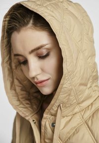 Beżowa, pikowana kurtka z kapturem i sznurkiem ściągającym, charakteryzująca się gładką teksturą oraz wzorem w diamenty na tkaninie.