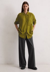 Grünes, oversized Hemd mit Kragen, ausgestattet mit einer einzelnen Brusttasche, kombiniert mit weiten, dunkelgrauen Hosen und schwarzen Schuhen.