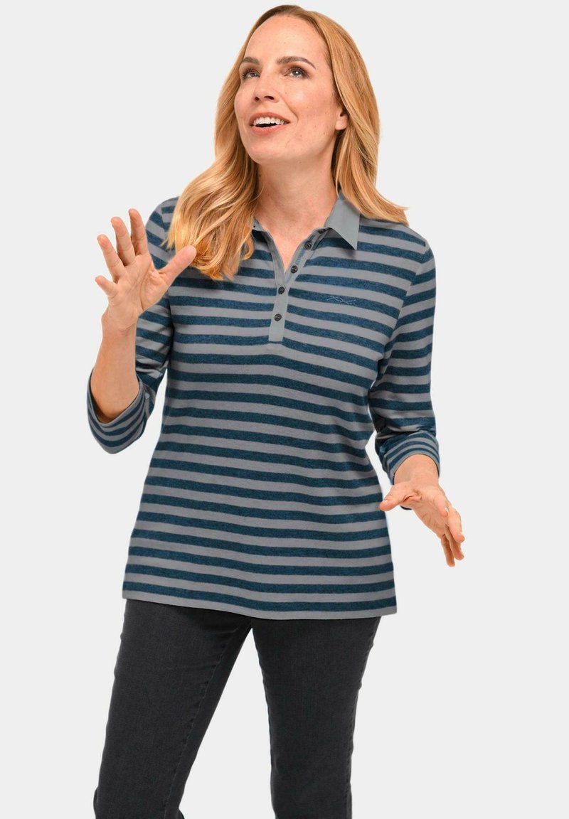 Gestreiftes Polohemd in Blau- und Grautönen mit drei Knöpfen; lange Ärmel und weiche Textur. Modell trägt es mit dunklen Jeans.
