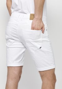 Shorts de mezclilla blanca con dobladillo enrollado, bolsillos traseros con solapas anguladas y una etiqueta de cuero en la cintura. Textura suave.