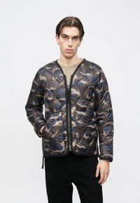 Fiatal, sötét hajú férfi karika fülbevalóval, barna és kék absztrakt mintás cipzáras steppelt dzsekiben, fekete nadrágban és bézs ingben.