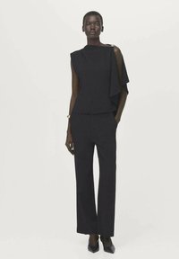Vrouw die een mouwloze zwarte top draagt met een gedrapeerd detail over één schouder en bijpassende zwarte wijde broekspijpen en puntige hakken.