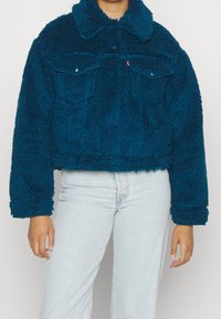 Teal, fluffig kort jacka med krage, två bröstfickor och långa ärmar. Mjuk textur och avslappnad passform. Minimalistisk design.