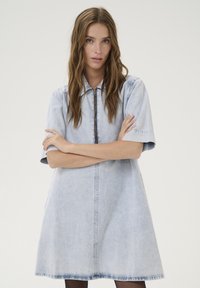 Robe chemise en denim bleu clair avec des manches courtes, fermeture éclair sur le devant et coupe décontractée. Présente une silhouette légèrement évasée.