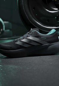 Chaussure de sport Adidas Primeweave noire et bleu sarcelle avec trois bandes argentées, présentée sur un fond de poids de salle de sport et de sol sombre.