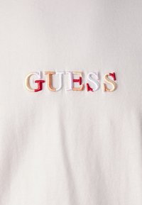 Camisa de color claro con el logo bordado de "GUESS" en hilos multicolores: rojo, melocotón, crema y lavanda sobre un fondo de tejido suave.