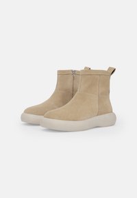 Beige suede nilkkurit, joissa on teksturoitu pinta, takaosan vetoketju ja valkoinen kumipohja. Sivussa vetoketjukiinnitys.