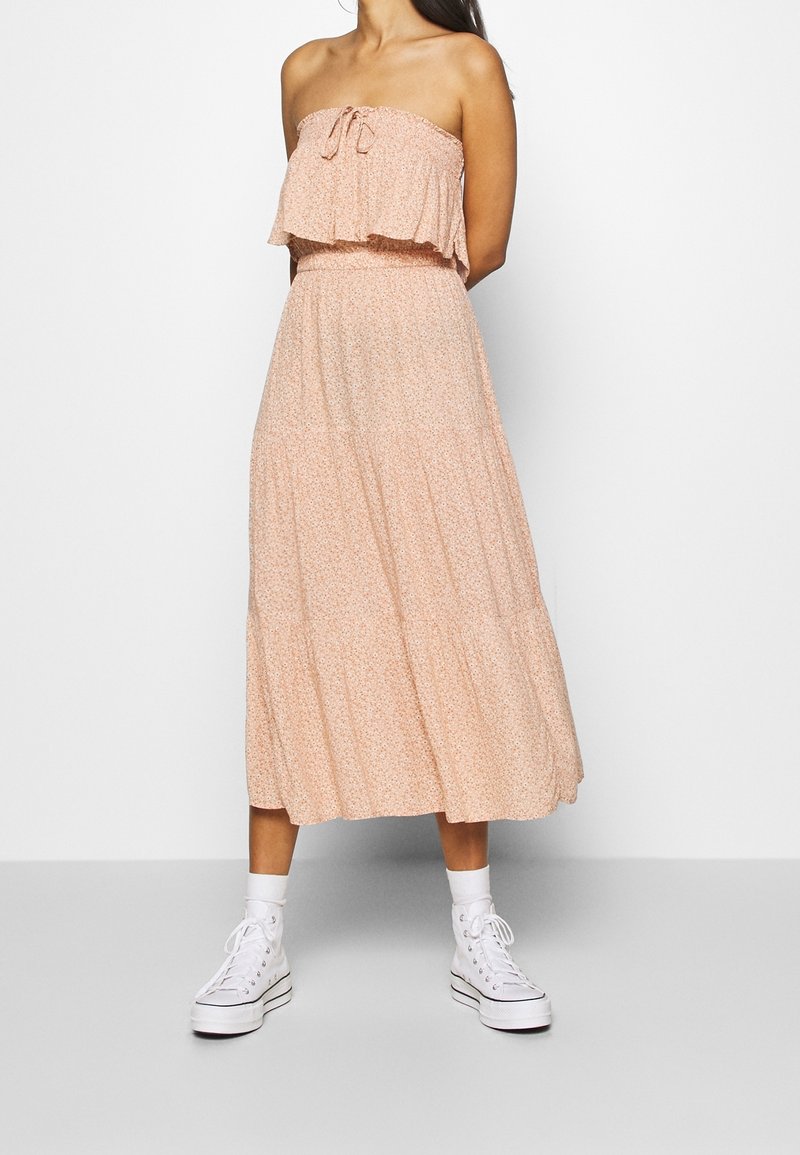 Robe maxi florale pêche avec un haut à volants superposés, une taille élastique et une jupe ample et fluide, associée à des baskets montantes blanches.