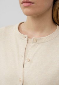 Beige cardigan met een ronde halslijn, ribbelstructuur en twee knoopsluitingen aan de voorkant. Gladde stof met subtiele variaties in toon.