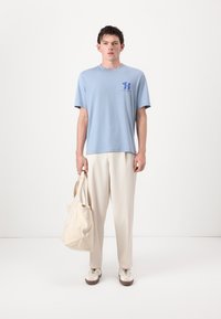 Lyseblå bomulls T-skjorte med korte ermer, blå logo på brystet, kombinert med kremfargede bukser og en beige handlenett. Hvite joggesko med brune såler.