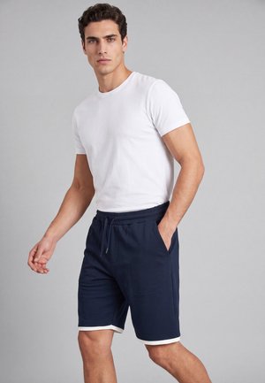 Uomo che indossa una maglietta bianca semplice e pantaloncini blu navy con bordino bianco, in piedi con una mano in tasca davanti a uno sfondo uniforme.