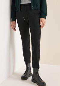 Černé tapered kalhoty vyrobené z texturované látky, s elastickým páskem na stažení. V kombinaci s černými šněrovacími bojovými botami.