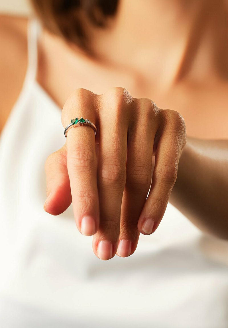 Hand med nakna naglar och en silverring med tre gröna stenar på långfingret, mot en suddig bakgrund av vit överdel.