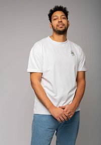 Weißes Baumwoll-T-Shirt mit hochgerollten Ärmeln, mit einem kleinen grünen Grafikdesign auf der Brust. Getragen mit hellblauen Jeans.