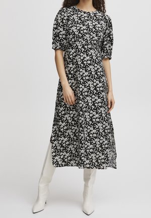 Kobieta w czarnej sukience midi z białym kwiatowym wzorem i bufkami na krótkich rękawach, zestawiona z białymi, szpiczastymi kozakami za kolano.