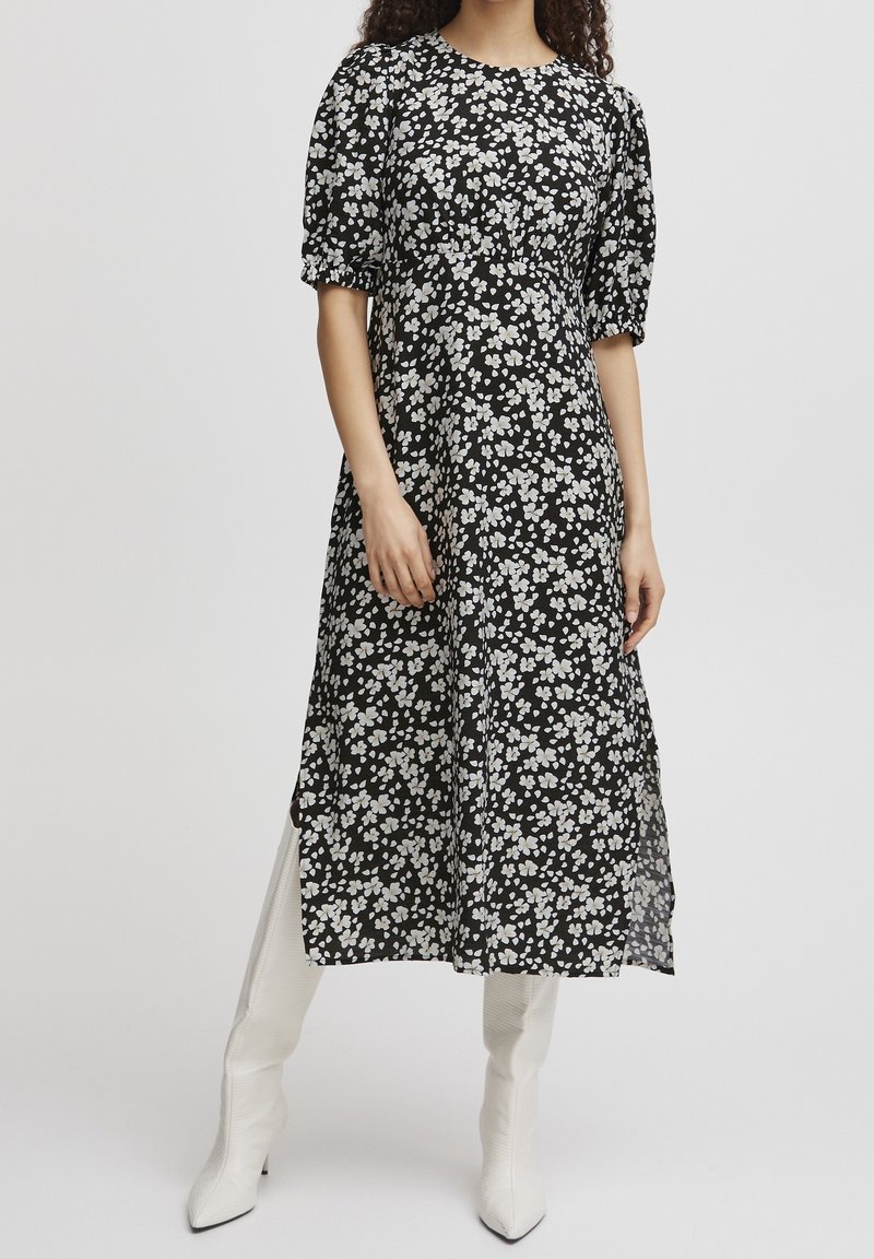 Femme portant une robe midi noire à imprimé floral blanc avec des manches courtes bouffantes, associée à des bottes blanches pointues montant au genou.
