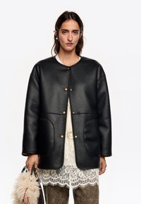Chaqueta de cuero negro con cuello redondeado, cierre de botones a presión, dos bolsillos laterales y textura suave, superpuesta a una blusa de encaje blanca.