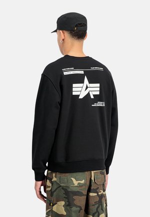 Sudadera negra con cuello redondo, que presenta un gran logotipo blanco y texto en la parte de atrás, combinada con pantalones cortos cargo de patrón de camuflaje.