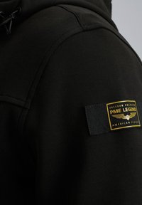 Sudadera negra con una textura suave, que presenta una etiqueta PMI Legend en dorado y negro, situada en el hombro izquierdo. Detalle con cordones visible.