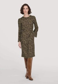 Leopardenmuster-Kleid in olivgrün, mit langen Ärmeln, Rundhalsausschnitt, knielang und seitlichen Taschen, kombiniert mit braunen knielangen Stiefeln mit Absatz.