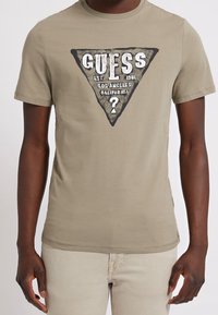 Camiseta de algodón verde oliva con un gráfico triangular en blanco y negro que incluye "GUESS" y detalles de la ciudad. Mangas cortas, cuello redondo, diseño ajustado.