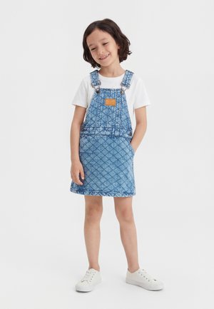 Χαμογελαστό παιδί που φοράει άσπρα αθλητικά παπούτσια, λευκό πουκάμισο με κοντά μανίκια και μπλε τζιν φόρεμα-σαλοπέτα με καπιτονέ μοτίβο, έχοντας τα χέρια στις τσέπες.