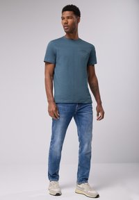 Blå crew neck t-shirt med en texturerad yta, ihopparad med ljusblå jeans. Modellen står i neutrala skor och vänder sig något uppåt.