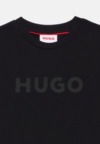 Μαύρο βαμβακερό μπλουζάκι με στρογγυλή λαιμόκοψη και έντονο, μονοχρωματικό λογότυπο "HUGO" στο μπροστινό μέρος. Κόκκινο στοιχείο στην περιοχή του γιακά.