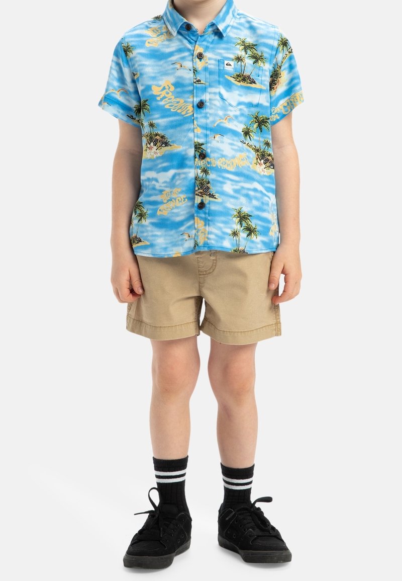 Enfant portant une chemise bleue à manches courtes à imprimé tropical, un short beige, des chaussettes noires avec des rayures blanches et des baskets noires, debout.