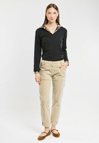 Donna in piedi con la mano in tasca, che indossa un top nero a maniche lunghe con colletto decorato e pantaloni beige con risvolto, scarpe basse marroni.