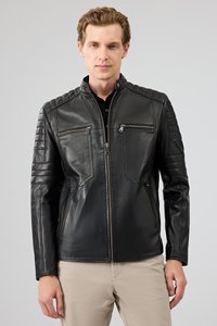 Man wearing a black quilted leather jacket with zippered pockets and beige pants, standing against a plain gray background.