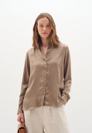 Camicia in satin color taupe con colletto rialzato e maniche lunghe; presenta un orlo curvo e un design minimalista. Pantaloni beige chiaro.