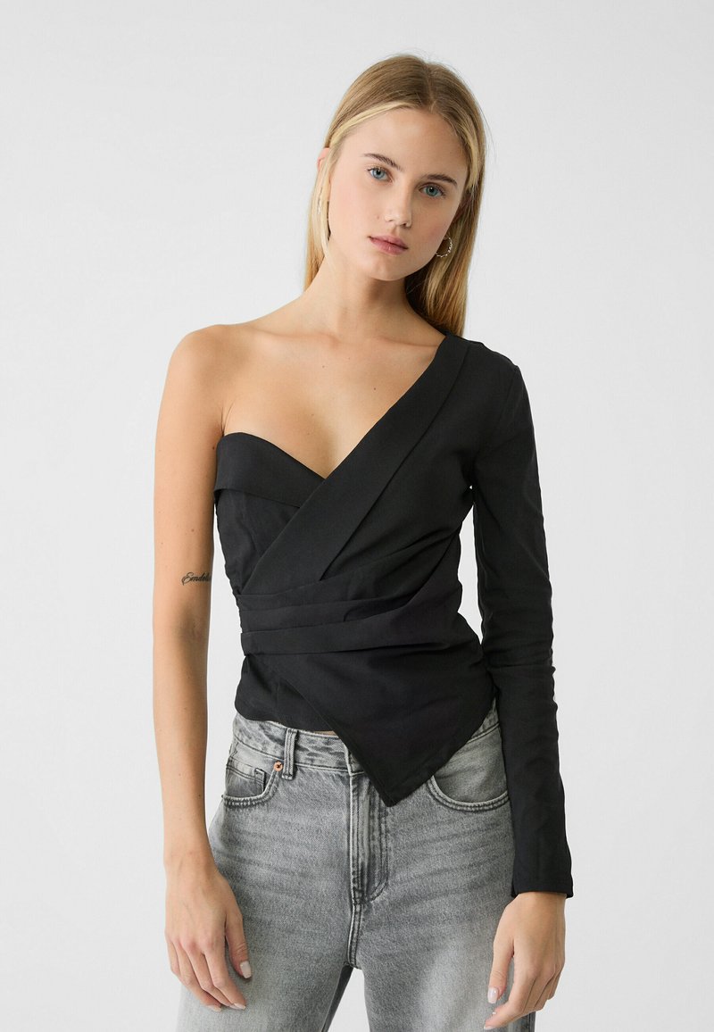 Top nero a spalla unica con design avvolgente, manica lunga da un lato, texture liscia e orlo asimmetrico, abbinato a jeans grigio chiaro.