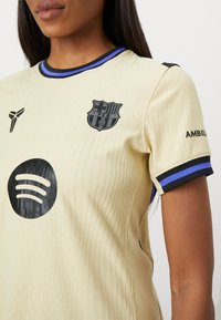 Krämfärgad sporttröja med texturerad tygkvalitet, korta ärmar med blå och svarta detaljer, samt logotyper för FC Barcelona och Spotify.