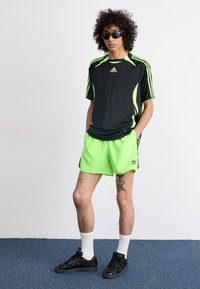 Tricou atletico negru cu accente verde neon, asortat cu pantaloni scurți verzi neon. Pantofi sport negri și șosete albe completează look-ul.