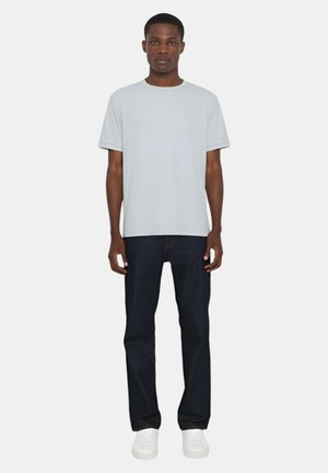 Maglietta grigia chiara a girocollo, maniche corte, vestibilità casual, abbinata a jeans blu scuro e sneakers bianche, in piedi davanti a uno sfondo neutro.