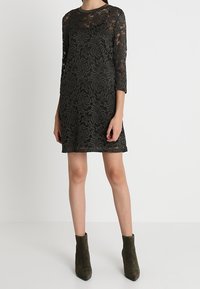 Svart spetsklänning med blommönster, 3/4 ärmar, rund halsringning och rak skärning, tillsammans med olivgröna ankelskor.