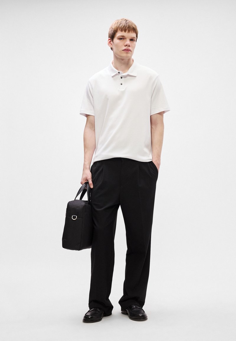Polo shirt blanc à manches courtes avec trois boutons, associé à un pantalon noir ample et des chaussures noires. Portant un petit sac noir.