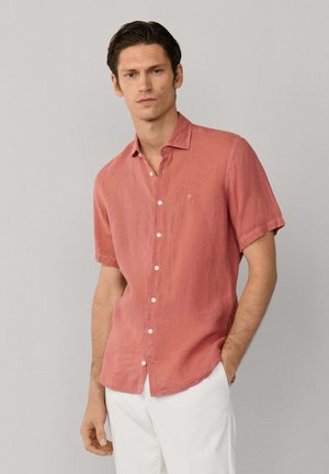 Hombre vistiendo una camisa terracota de manga corta y botones, con pantalones blancos, posando frente a un fondo liso de color gris claro.