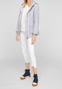 Veste à capuche gris clair avec fermeture éclair et poches, portée sur un haut blanc, associée à un pantalon blanc court et des baskets montantes bleu marine.