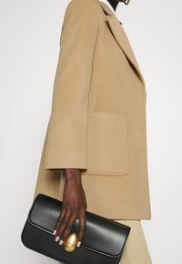 Person in beigem Mantel hält eine schwarze Clutch mit goldener Schnalle, zeigt Handgelenk und Hand mit neutralem Nagellack vor weißem Hintergrund.