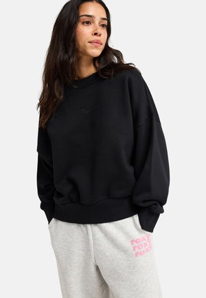 Frau mit langen dunklen Haaren, die einen lockeren schwarzen Sweatshirt und hellgraue Jogginghose mit pinkem "roxy" Schriftzug am linken Oberschenkel trägt.