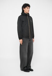 Vyras stovintis šone, vilkintis juodą užtrauktuku užsisegamą gobtuvą, plačius pilkus džinsus ir juodus batus prieš baltą foną.